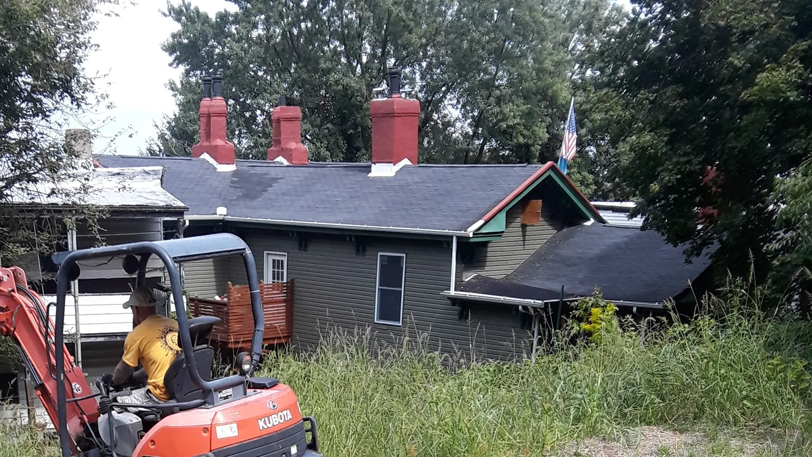 Demolition contractor serving Plum, PA: operator working a mini excavator in an overgrown lot beside a gray house with red brick chimneys
